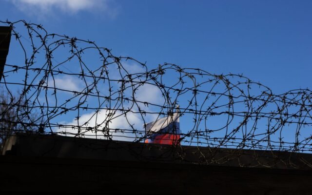 Silhouette of barbed wire with a flag in the background against a clear blue sky