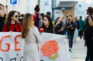 Journalist interviews protesters during a city rally with people holding banners and flags