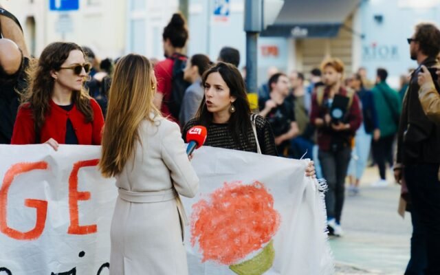Journalist interviews protesters during a city rally with people holding banners and flags