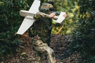 A soldier in camouflage gear carrying a drone in a dense forest setting