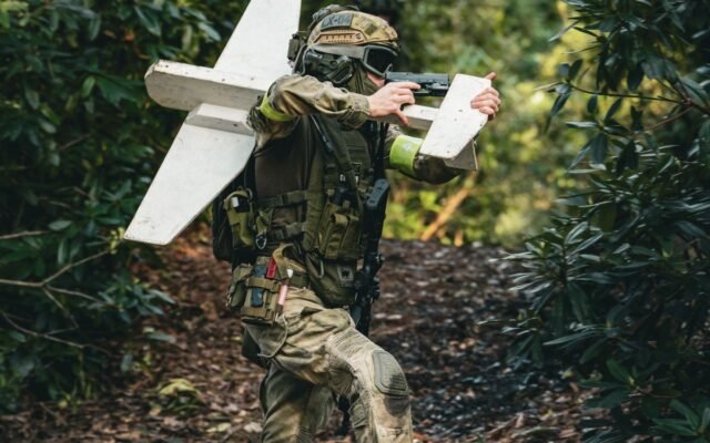 A soldier in camouflage gear carrying a drone in a dense forest setting